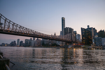The Queensboro Bridge spans across the East River as Manhattan's illuminated skyline reflects in the calm water. The scene captures the peaceful beauty of the city during the last light of day.