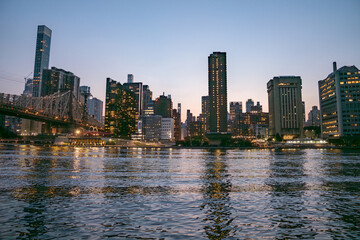 The Manhattan skyline and Queensboro Bridge glow with city lights, mirrored in the rippling East River. The sky transitions from soft blue to warm tones as the city settles into night.