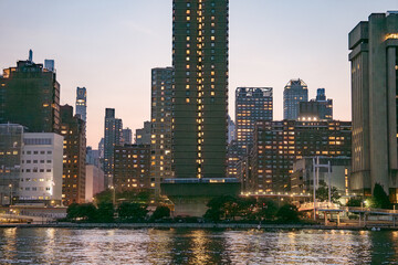 Tall residential buildings in Manhattan glow with warm lights at twilight, reflecting beautifully on the East River. The scene captures the city's calm, vibrant atmosphere as evening settles in.