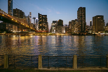 The New York City skyline glows in the evening light with skyscrapers, a lit bridge, and shimmering reflections on the river. A black metal fence lines the quiet waterfront, contrasting with the