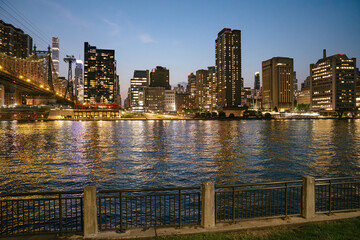 The New York City skyline shines with illuminated skyscrapers beside the Queensboro Bridge at twilight. The river reflects golden lights, and a quiet metal fence lines the calm waterfront in the