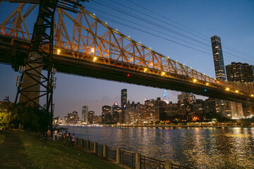 The Queensboro Bridge stretches over the East River as the Manhattan skyline glows in the background. A group of people stands by the waterfront, enjoying the city lights and evening atmosphere.