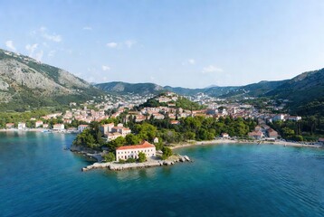 Obraz premium Coastal charm: aerial view of a picturesque town on a peninsula surrounded by serene blue sea and lush mountains.