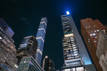 Tall skyscrapers rise sharply into the night sky in Midtown Manhattan, their windows glowing against the dark. A striking architectural view captures the vertical energy and modern power of New York