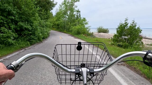 POV Lakeside Bike Ride. Slow-motion POV of a cruiser bike ride on a paved coastal road. Shows handlebars and basket with dense green forest on one side and sparkling blue lake waters on the other.
