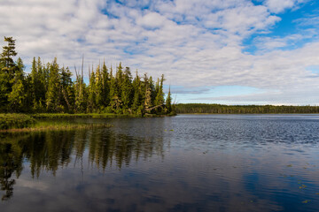 Obraz premium Vancouver Island's Keta Lake’s calm surface reflecting a partly cloudy sky and blue light, framed by evergreen shoreline