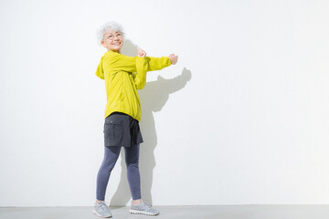 白背景でスポーツウェアでストレッチで腕を伸ばすグレイヘアのシニア女性の全身