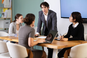 Fototapeta premium Confident young businessman standing near table where sitting colleagues talking and discussing ideas during business meeting in bright office