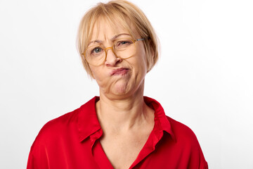 Fototapeta premium Mature woman with short blonde hair wearing glasses and red shirt makes a skeptical face against white background. Concept of doubt, confusion, and mixed emotions in portrait photography.