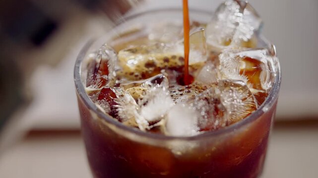 Close up of iced coffee pouring over ice cubes in glass. Refreshing cold caffeine beverage with bubbles and splash, perfect for summer drink and cafe menu concept.