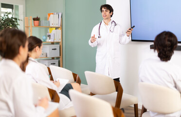 Fototapeta premium Young male doctor in medical gown with remote control gives presentation to colleagues doctors in conference room