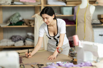 Fototapeta premium Dedicated young female dress designer working in creative fashion studio, developing new patterns, drawing and adjusting layout for future garment on paper using curve ruler