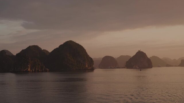 Warm sunset light illuminates calm waters and layered limestone karst silhouettes in Ha Long Bay, Vietnam, creating a tranquil and atmospheric seascape at dusk.