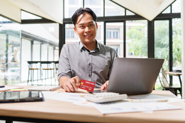 Obraz premium A man is sitting at a desk with a laptop and a stack of papers. He is smiling and he is enjoying his work. The desk is cluttered with various items, including a keyboard, a mouse, and a few books 