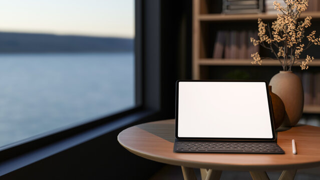 White screen tablet with keyboard and stylus on round wooden table aside bookshelf and glass window.
