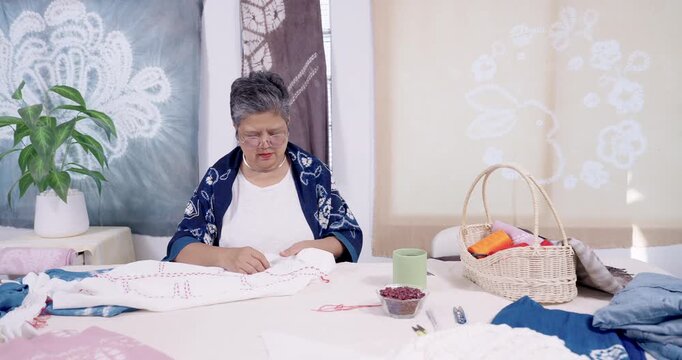 Senior Asian woman artisan stitches resist pattern on cotton cloth, running family small business in home studio, guiding slow craft shibori tie dye preparation step by step for sale, heritage.