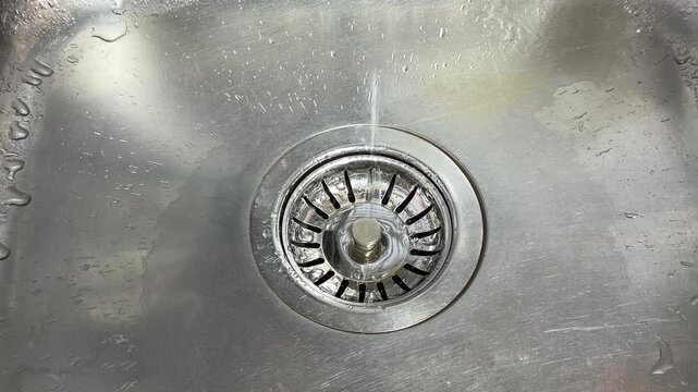 A metal mesh stopper sits inside a stainless steel kitchen sink drain. Residual water droplets cling to the scratched metal surface, creating a faint, rhythmic "plink" as they hit the drain.