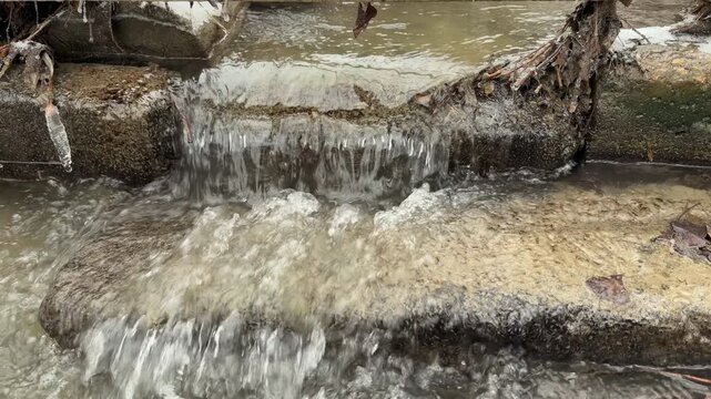 Water flows steadily over a concrete ledge and mossy stones in a snowy forest. This continuous movement against the landscape creates a low-frequency hum and a rhythmic, liquid gurgling sound.