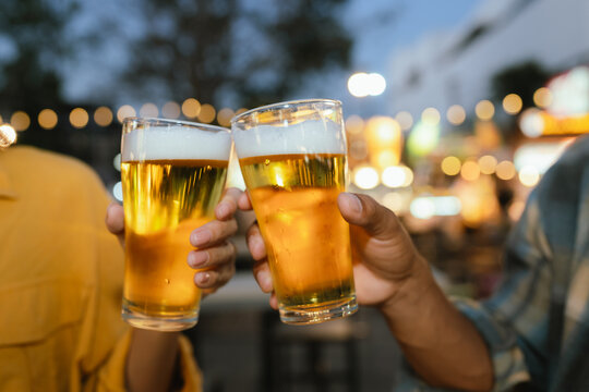 Two people clinking glasses of cold beer at an outdoor night market, celebrating with street food and evening lights in the background.