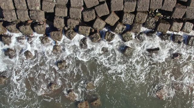 Drone footage of sea waves crashing against square concrete wave breakers on the shore. Industrial coastal protection and water textures from above. 4K. Real time.