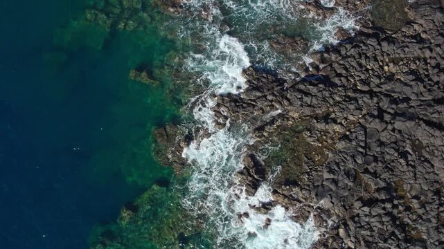 Drone shot of turquoise sea water splashing against a rugged black rocky shoreline. Clear ocean textures and natural coastal beauty from above. 4K. Real time.