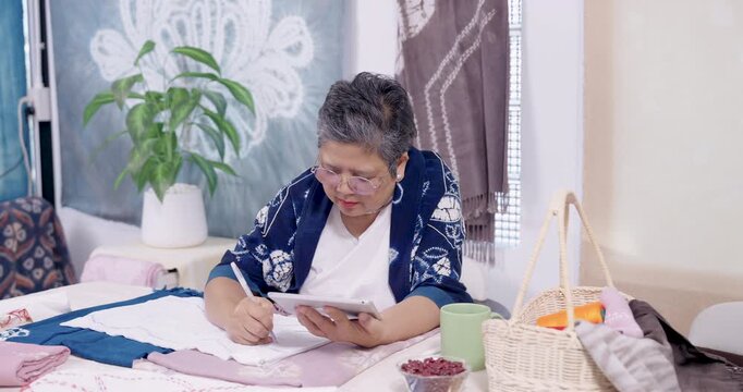 Retired Asian woman entrepreneur swipes tablet, checks design note, then marks fabric pattern line, coordinating shibori resist stitching plan for indigo dye, heritage slow fashion textile order.