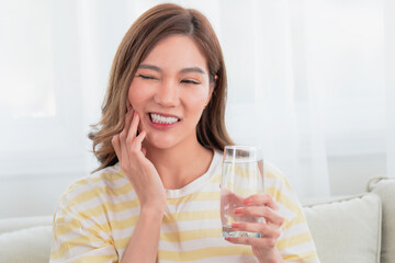 Young Asian woman with a pained expression, touching her cheek while holding a glass of water. Concept of sensitive teeth, toothache, and oral hygiene.