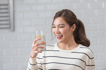 Happy young Asian woman in a striped sweater holding a glass of water with an orange slice in a bright kitchen. Concept of healthy hydration and vitamin C.