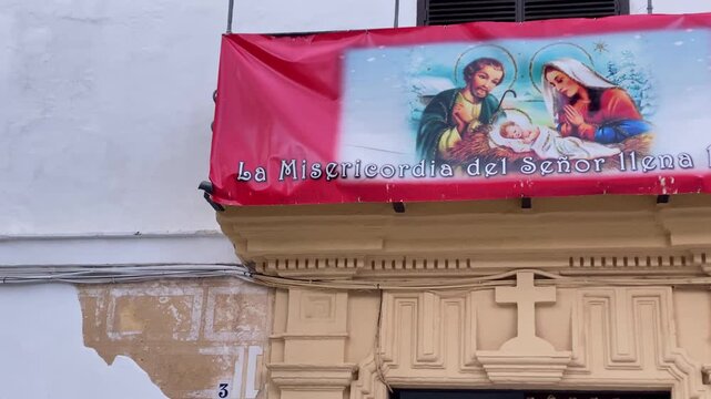 Public street sign for Plaza de Santa Angela de la Cruz on a weathered white building wall with a saint portraits