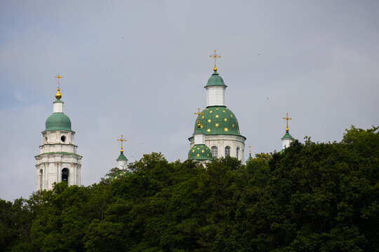 The beautiful architecture of the Mgarsky Spaso-Preobrazhensky Monastery with its green and gold decorated domes. The church steeples are visible behind a lush green forest under a cloudy sky.
