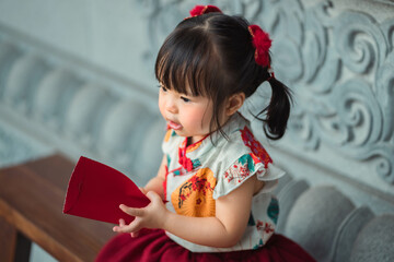 Closeup - Adorable little Asian girl in traditional Chinese dress holding red envelope angpao, sitting on wooden bench at shrine, celebrating Chinese New Year festival, Lunar new year
