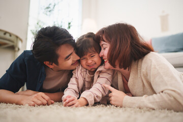 Happy Asian parents kissing and playing with daughter on carpet at home