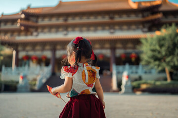 Back view of little Asian girl in traditional Chinese dress holding red envelope at temple, cute toddler looking at beautiful oriental architecture during Lunar New Year festival, celebration.