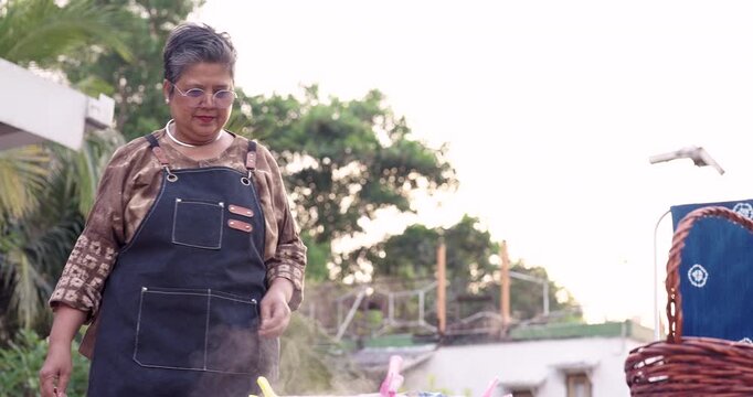 Senior Asian woman entrepreneur clips cloth and secures bundle at dye station, preparing resist tied fabric for indigo dyeing, outdoor home studio workflow for handmade textile customer order.