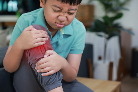 Asian boy showing painful face expression holding red inflamed knee from joint injury or accident. Pediatric healthcare concept of childhood bone problems, growth pain, and orthopedic treatment.