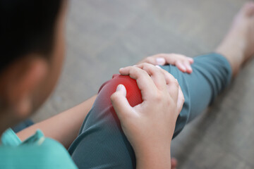 Naklejka premium Little boy holding red inflamed knee with hands suffering from joint pain or accidental injury at home. Concept of pediatric healthcare, bone problems in children, and orthopedic medical treatment.