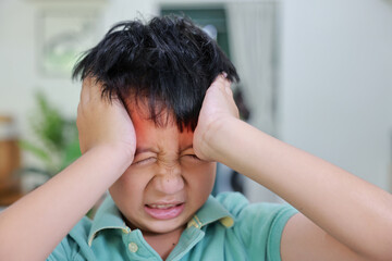 Fototapeta premium Asian boy suffering from severe headache or migraine with red highlight on forehead, child having fever, dizziness, brain tumor symptoms or stress while touching head at home.
