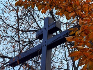 Wooden cross against autumn leaves and bare tree branches.