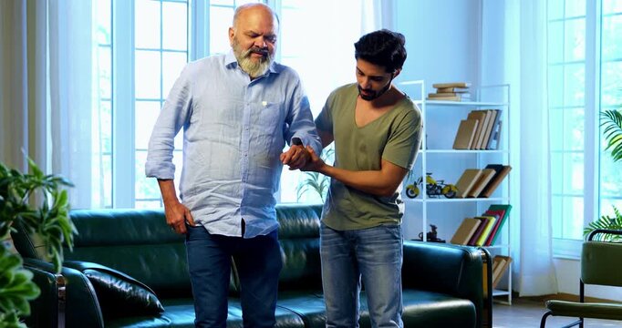 Indian young son helping and assisting aged father stand and walk with his physical support in living room at modern home, showing patience, strength and responsibility toward elderly parent