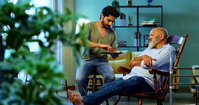 Indian young son feeding senior father chapati with sabzi from plate while he relaxes on chair, sharing heartfelt conversation and affection in modern home living room during homely mealtime