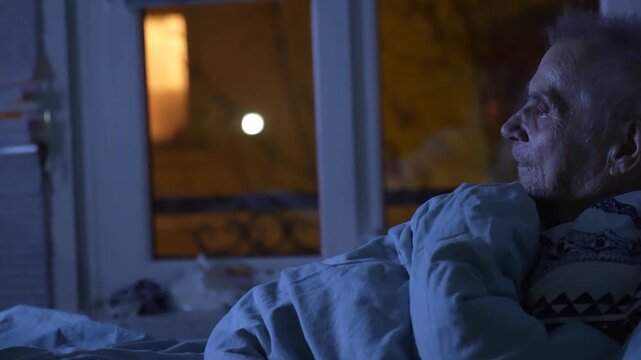 Senior Man Lying in Bed Watching Television at Night. Elderly man resting in bed in a dimly lit room, looking at a bright TV screen. Concept of senior lifestyle, aging, loneliness
