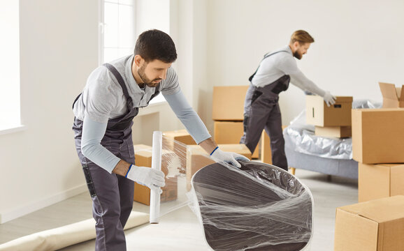 Two professional movers in uniform pack furniture and cardboard boxes during a home relocation. One wraps a chair while the other boxes. A moving service provides efficient cargo transportation. 