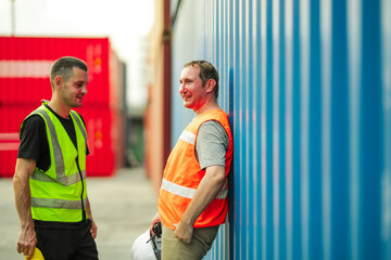 Two industrial workers talking beside blue shipping container at busy cargo yard. They discuss operations and logistics plans while wearing safety vests and hard hats in global trade environment.