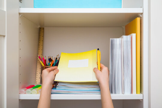 Little child hands holding copybook and pen. Different school accessories on white wardrobe shelf. Closeup. Making neatness. Point of view shot. Front view.
