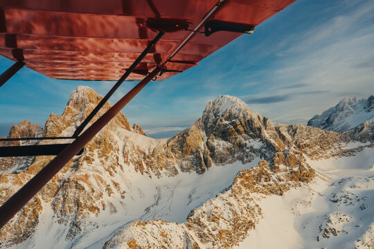 Red airplane wing flys across snow covered alpine ridge at sunset.