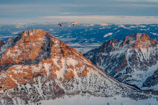 Small airplanes fly past snow covered mountain peaks.