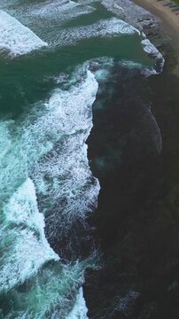 Aerial drone footage shows the dramatic rocky coastline and ocean currents flowing along at Sawarna Beach, Indonesia. The powerful natural composition is presented in a high-quality vertical frame.