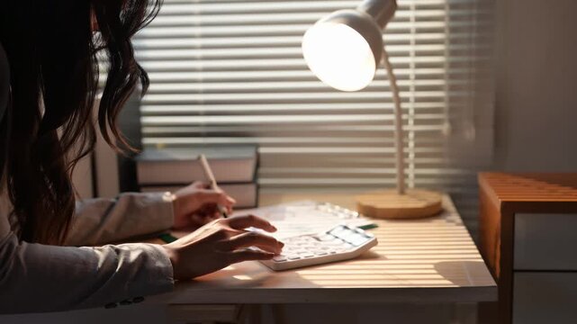 Businesswoman using calculator and taking notes under a desk lamp