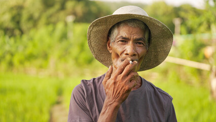 Portrait, old indonesian man farmer smoking a cigarette in rural indonesia in agricultural field or farm, looking at camera, wearing straw hat, southeast asia