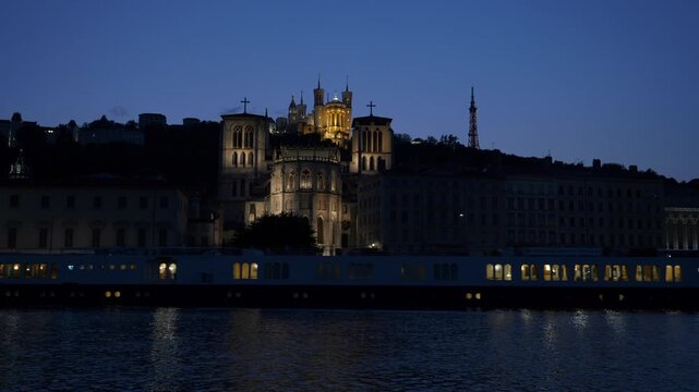 Basilique de Fourvi&egrave;re illumin&eacute;e au cr&eacute;puscule &agrave; Lyon avec bateau mouche sur la Sa&ocirc;ne, paysage urbain nocturne, architecture historique et lumi&egrave;res de la ville en ambiance calme et cin&eacute;matographique